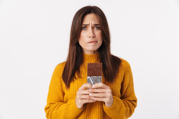 Image of beautiful brunette adult woman hesitating while holding chocolate bar isolated on white