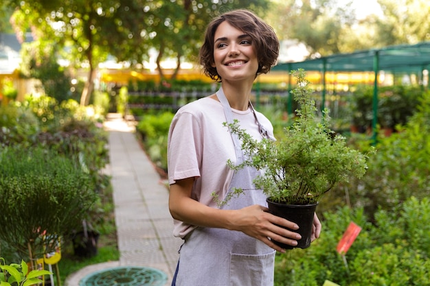 Image of beautiful cute woman gardener standing over flowers plants in greenhouse holding plants