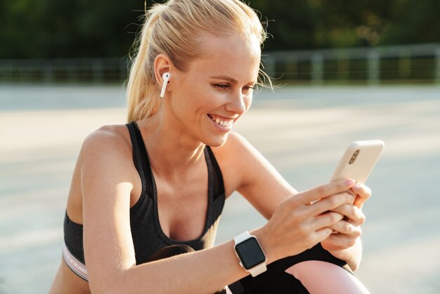 Image of beautiful sportswoman wearing tracksuit and earpods holding cellphone while doing workout outdoors