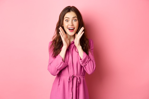 Image of beautiful surprised woman gasping amazed, look at fantastic news, smiling astonished, standing over pink wall.