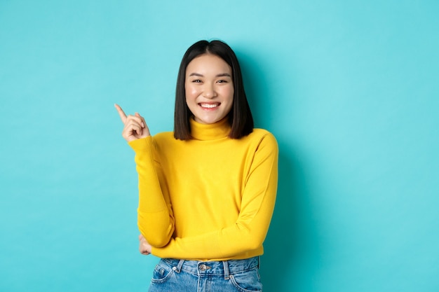 Image of beautiful young asian woman with perfect smile and no blemishes on skin, pointing finger left, looking satisfied, standing over blue background.