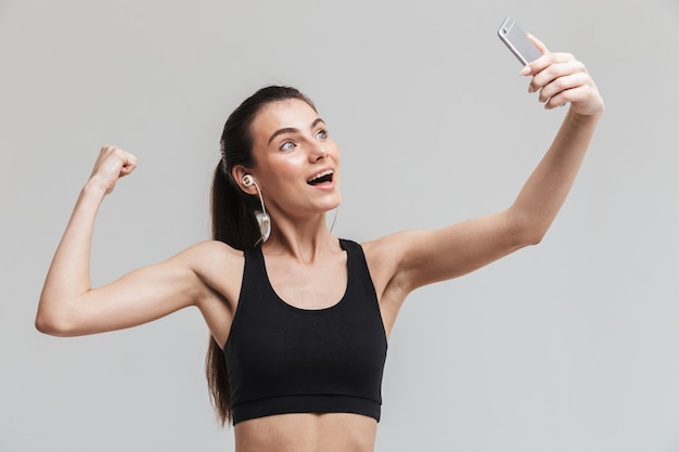 Image of a beautiful young sport fitness woman using mobile phone listening music isolated over grey wall take a selfie showing biceps.