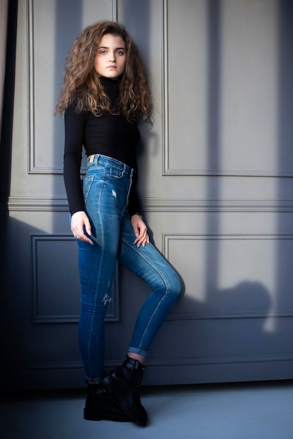 Image of a beautiful young woman with curly hair in black suitcase and jeans posing near grey wall