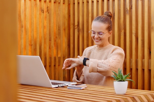 Image of busy happy young adult woman with bun hairstyle wearing beige sweater working on notebook and using smartwatch looking at gadget with smile in office against wooden wall