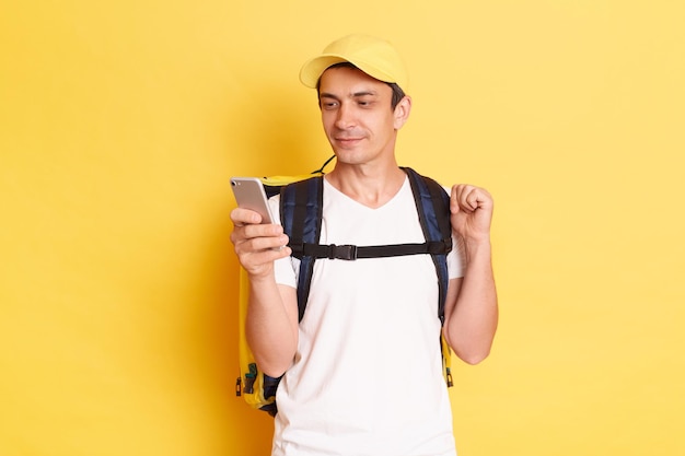 Image of calm concentrated courier man in yellow cap and tshirt holding smart phone and looking at screen finding address of client surfing internet during break isolated on yellow background