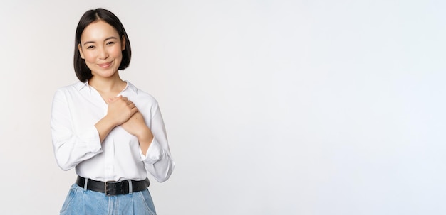 Image of caring young beautiful asian woman holding hands on heart looking pleased and happy smiling