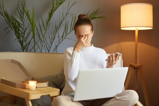 Image of Caucasian exhausted unhealthy woman wearing white sweater sitting on sofa and working on laptop holding smart phone in hands rubbing her eyes