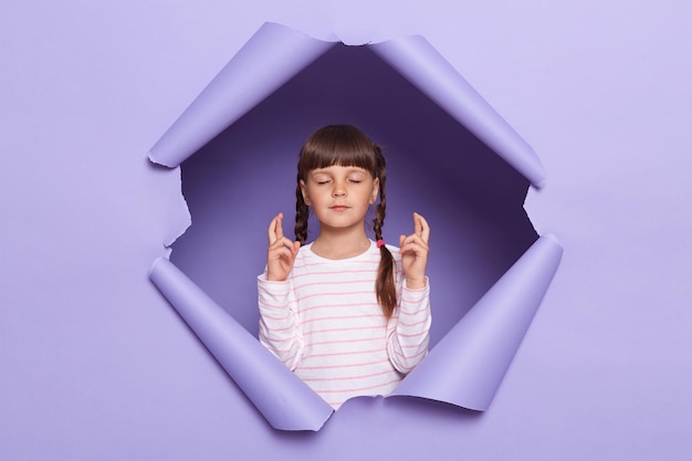 Image of charming little girl with braids wearing striped shirt posing in torn paper wall standing with closed eyes and crossed fingers making wish dreaming of the best