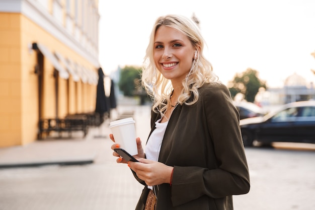 Image of charming woman wearing earphones holding takeaway coffee and mobile phone, while walking through city street