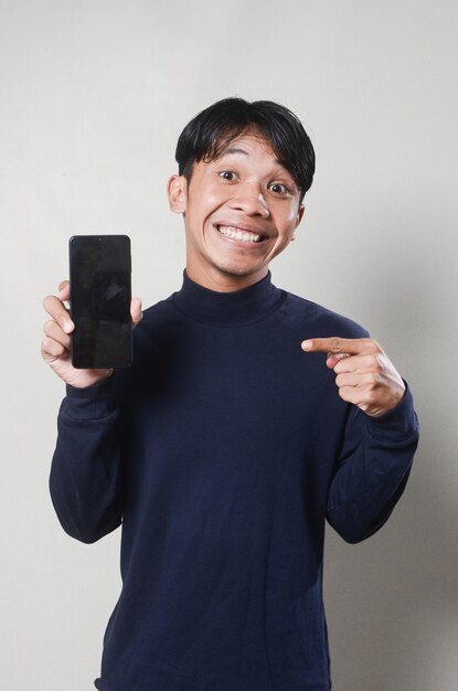 Image of cheerful asian man smiling and pointing at mobile phone