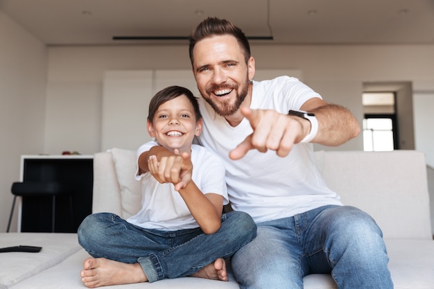 Image of cheerful glad father and son laughing, while pointing finger at you and sitting on sofa in apartment