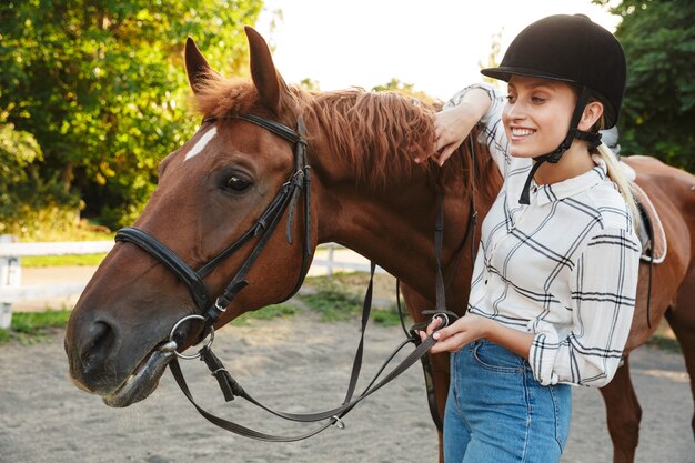 Image of cheerful young blonde woman wearing hat standing by horse at yard in countryside