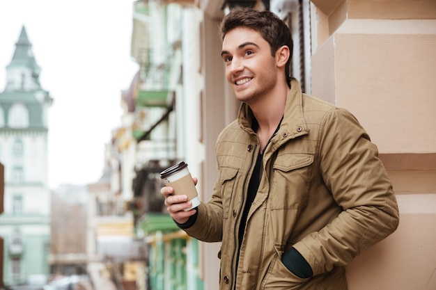 Image of cheerful young man walking on the street and looking aside while holding cup of coffee.