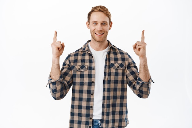 Image of confident redhead man pointing fingers up and smiling happy introduce company logo showing advertisement promo offer standing over white background