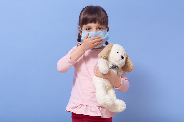 Image of cute European girl of preschool age dresses medical mask and holding dog toy in hand, cover mouth with palm