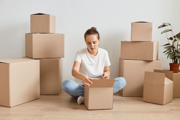 Image of dark haired girl in jeans and white tshirt unpacking cardboard box attractive european woman packing things to move packed boxes on the floor relocation and moving to new apartment