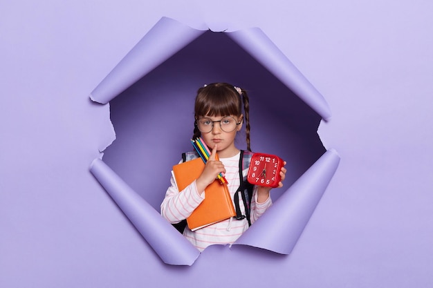 Image of dark haired little girl in a striped shirt with books and alarm clock in her hands and a backpack posing isolated over lilac background looking at camera with serious expression