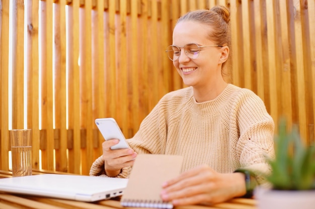 Image of dark haired positive woman with bun hairstyle wearing beige sweater holding paper organizer and cell phone looking smiling at display expressing happiness