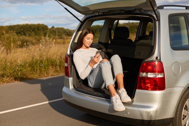 Image of dark haired young adult woman traveler sitting in car trunk and using cell phone female wearing casual clothes surfing internet checking social networks while traveling