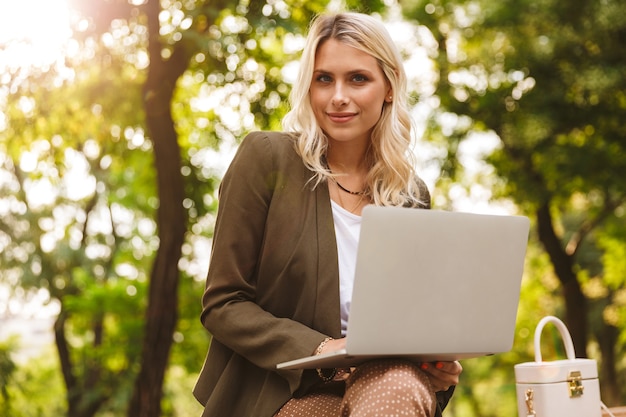 Image of elegant woman 20s using silver laptop, while sitting on bench in park