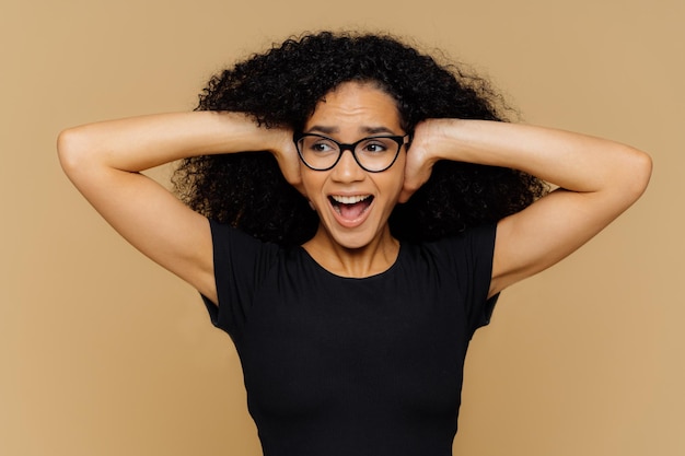 Image of emotional African American woman covers ears yells loudly cant stand noise demands turn off music dressed in casual black t shirt optical eyewear isolated over brown background