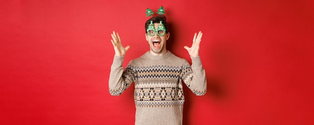 Image of excited and amazed caucasian guy in party glasses christmas sweater raising hands up and ma...