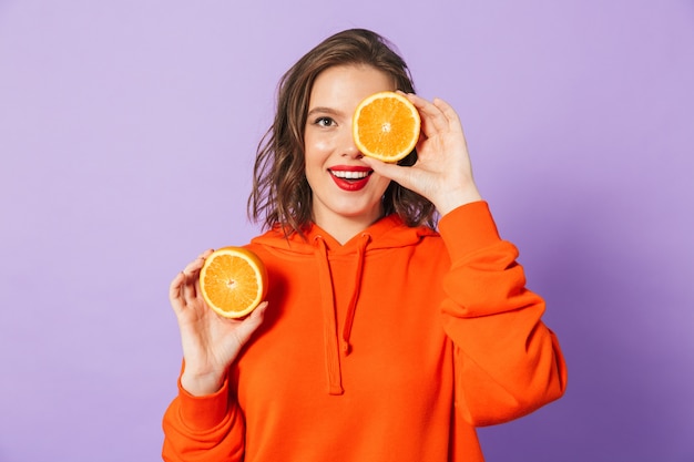 Image of an excited beautiful young woman posing isolated over purple wall wall holding orange citrus covering eyes.