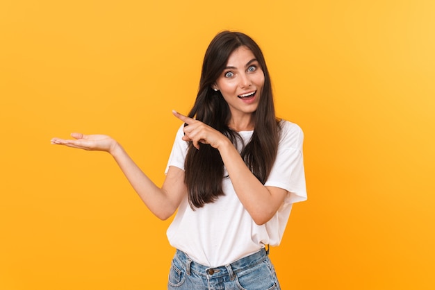 Image of excited brunette woman with long hair smiling and pointing fingers at copyspace on her palm isolated over yellow wall