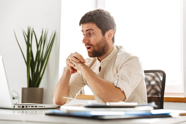 Image of excited businessman 30s wearing white shirt working with laptop and paper documents, while sitting in modern office