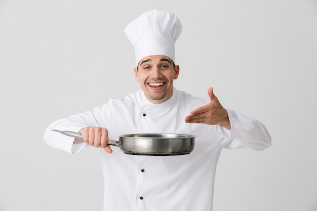 Image of excited emotional young man chef indoors isolated over white wall background holding frying pan.