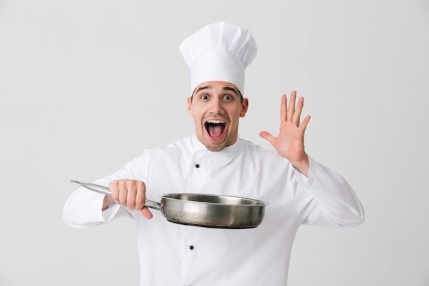 Image of excited emotional young man chef indoors isolated over white wall background holding frying pan.