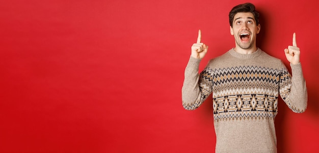 Image of excited handsome guy who likes winter holidays showing advertisement looking and pointing fingers up with amazed face standing in christmas sweater over red background