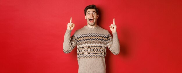 Image of excited handsome guy who likes winter holidays showing advertisement, looking and pointing fingers up with amazed face, standing in christmas sweater over red background