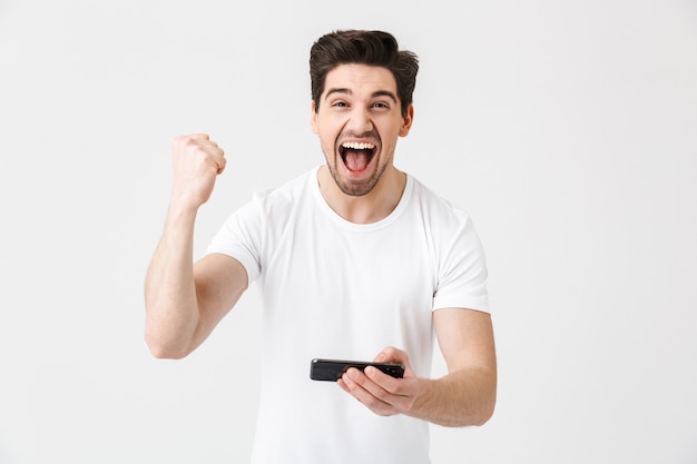 Image of excited happy emotional young man posing isolated over white wall  play games by mobile phone make winner gesture.