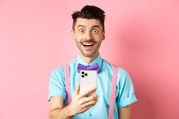 Image of excited man checking out promo offer online, holding smartphone and stare surprised at camera, standing happy on pink.