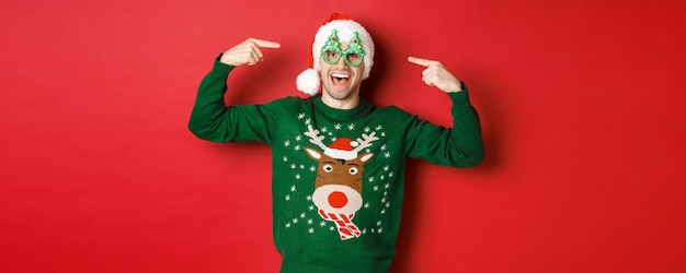 Image of excited smiling guy pointing at party glasses and celebrating new year standing joyful in g...