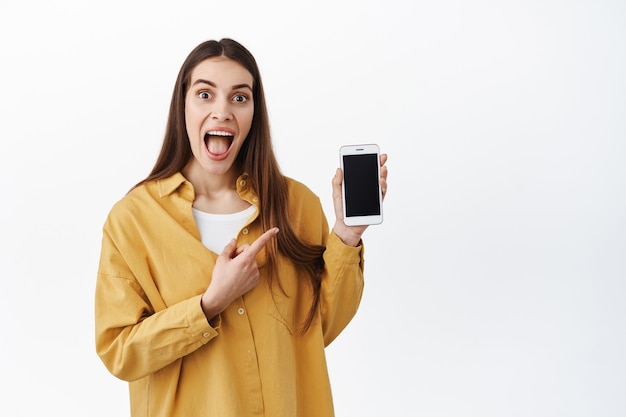 Image of excited woman checking out cool new online promo, pointing at smartphone and scream from joy and amazement, showing cool internet shop or application, standing over white wall