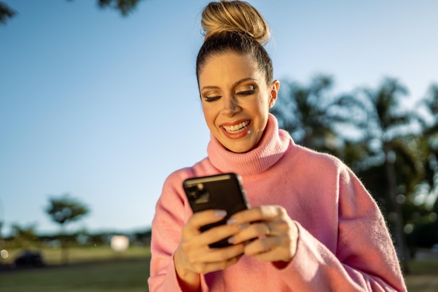 Image of excited young blond Latin woman happy using cellphone on park background