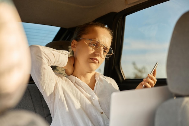 Image of exhausted business woman working laptop in car holding smart phone in hand looking at display of notebook feels pain in neck sitting in one position long hours