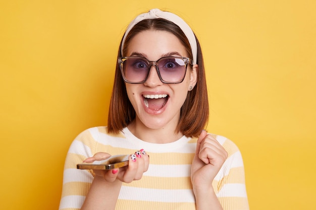 Image of exited positive woman wearing striped t shirt and sunglasses posing isolated over yellow background standing cell phone and clenched fist betting and winning celebrating her success