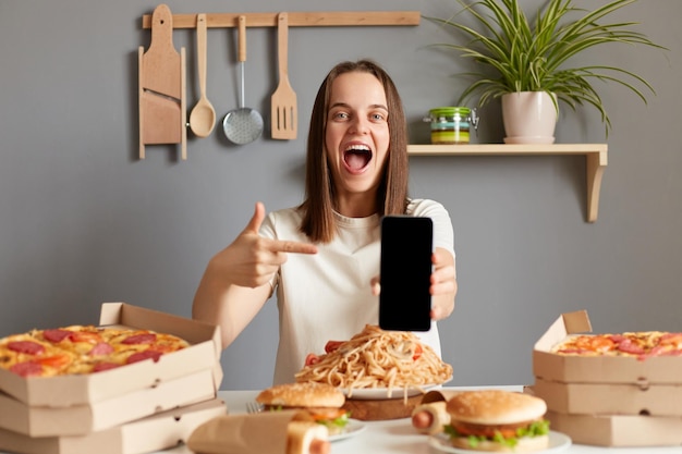 Image of extremely happy overjoyed woman with brown hair wearing white Tshirt sitting at table in kitchen pointing at mobile phone with black blank screen copy space screaming with happiness