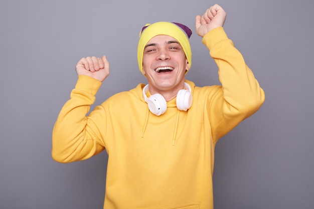 Image of festive happy cheerful man wearing beanie hat and yellow casual hoodie standing isolated over gray background posing with raised arms dancing and laughing celebrating