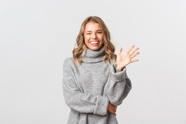 Image of friendly happy woman in grey sweater saying hello, smiling and waving hand to greet person