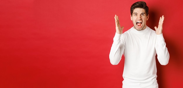 Image of frustrated and angry man in white sweater, shouting in rage, being mad at someone, standing over red background.