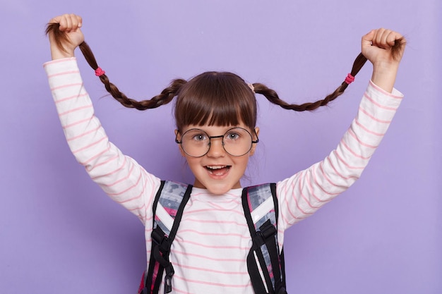 Image of funny pretty little girl with pigtails wearing backpack standing isolated over purple background raised her braids screaming looking at camera