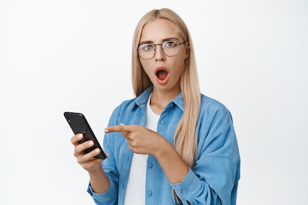 Image of girl in glasses holds phone points finger at smartphone with surprised and shocked face stands against white background