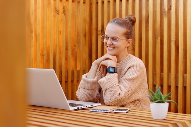 Image of good looking beautiful Caucasian woman with bun hairstyle wearing beige jumper and glasses working on notebook against wooden wall keeps hand together watching something pleasant on laptop