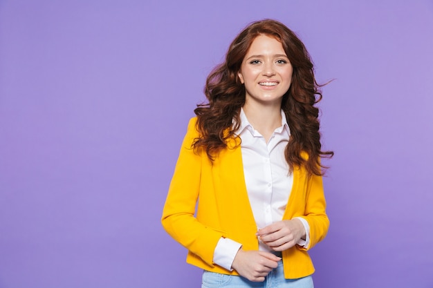 Image of gorgeous redhead woman wearing yellow jacket smiling and looking at camera over purple