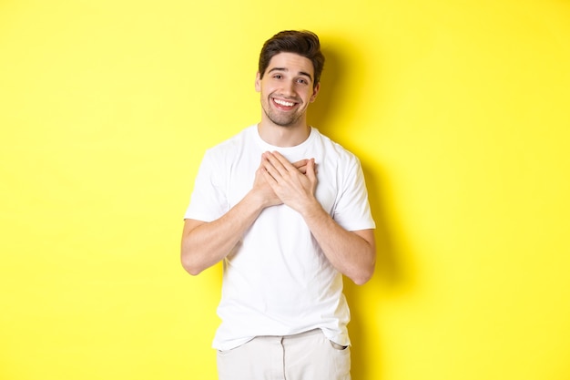 Image of grateful handsome guy in white tshirt holding hands on heart and smiling pleased express gr...