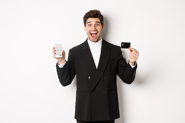 Image of handsome businessman in black suit, looking excited and showing credit card with mobile phone screen, standing against white background